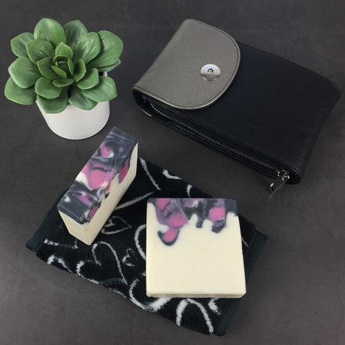 Black Raspberry Artisan Soap drop swirl design in raspberry, black, and white on slate background with a small black purse, houseplant, and beauty towel